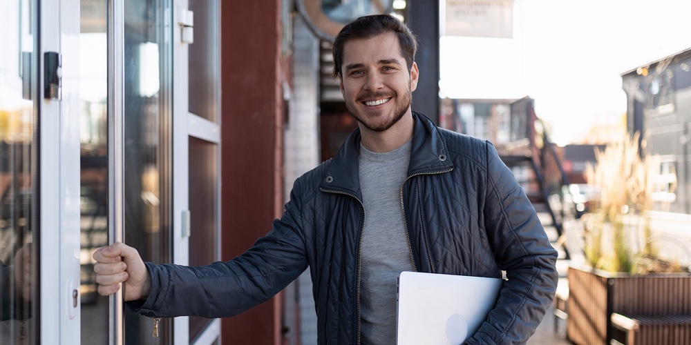 A Male It Specialist With A Laptop In His Hands Enters The Office Building With A Wide Smile On His