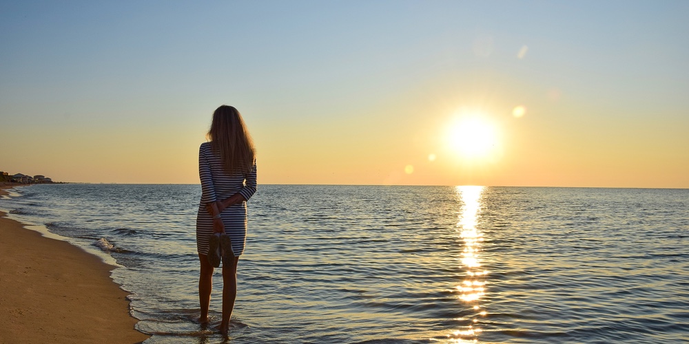 A Young Woman Is Walking Along A Deserted Sea Beach On The Shore Holding Her Shoes In Her Hands. Bac