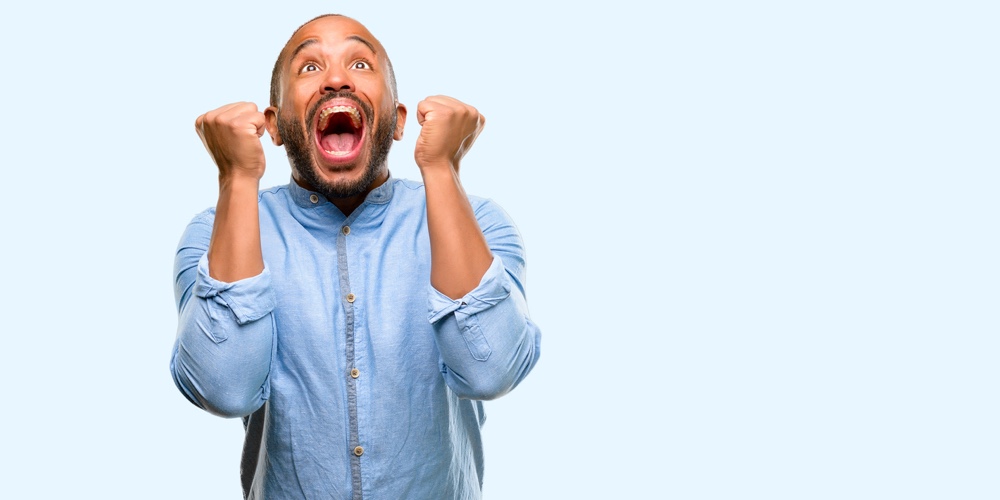 African american man with beard happy and surprised cheering expressing wow gesture isolated over bl