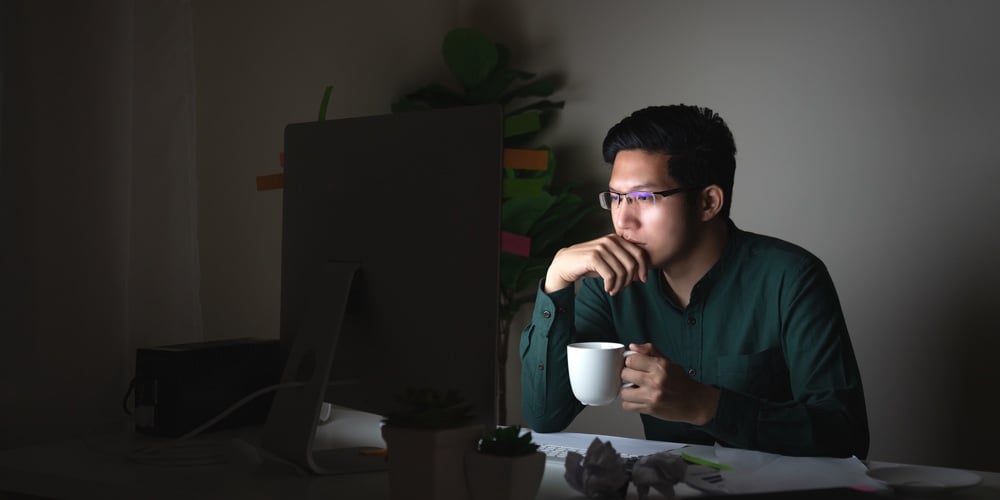 Attractive Young Asian Man Drinking Coffee Sitting On Desk Table Looking At Laptop Computer In Dark