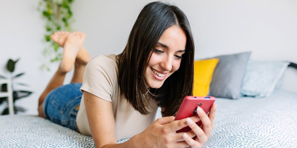 Beautiful Young Woman Holding Smart Phone And Looking At Smart Phone With Smile While Sitting In Com|Beautiful Young Woman Using Mobile Phone While Relaxing At Home. Smiling Pretty Girl Lying On Bed Lo|Young Woman Smiling Happy While Texting On Her Mobile Phone