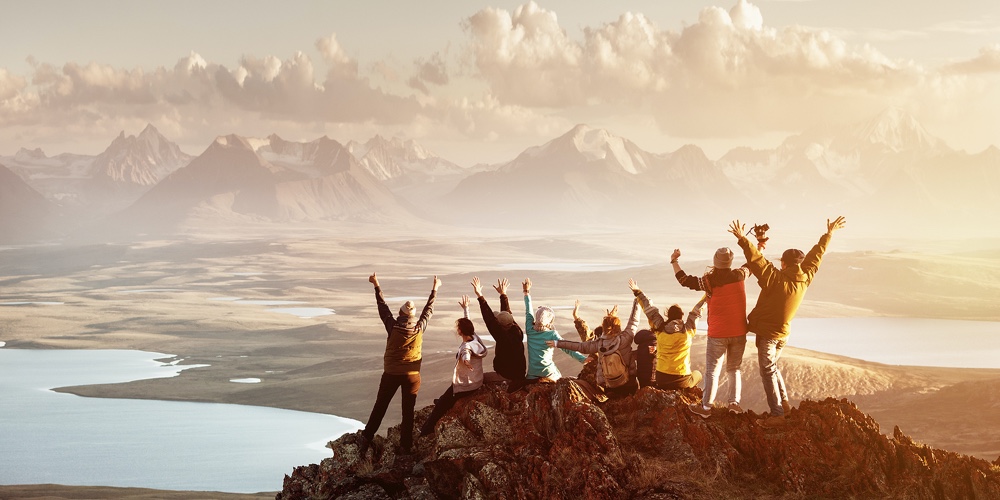 Big Group Of People Having Fun In Success Pose With Raised Arms On Mountain Top Against Sunset Lakes