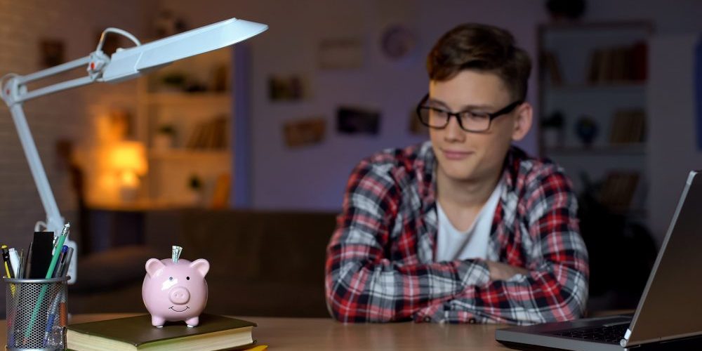 Cheerful Teenager Looking Into Money In Piggy Bank