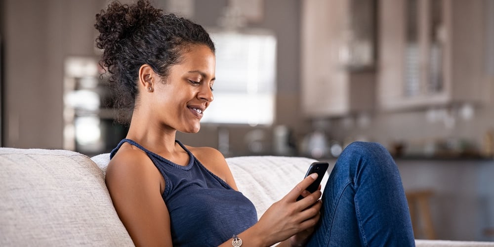 Cheerful young african woman using smartphone while sitting on couch. Black smiling woman using app
