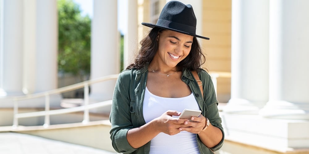 Cheerful young woman wearing black hat in city street typing a message. Hispanic trendy girl browsin