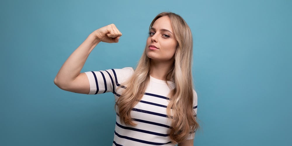 Close-up Photo Of A Strong Girl In Casual Clothes Showing Biceps On A Blue Isolated Background