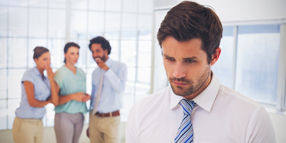 Colleagues gossiping with sad young businessman in foreground at a bright office