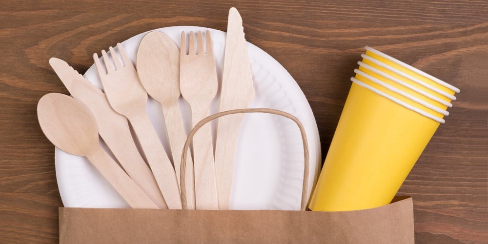 Disposable paper plates and cups and wooden cutlery on wooden table in a paper bag