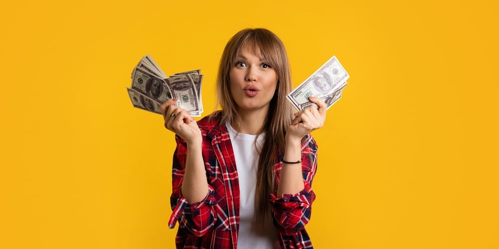 Excited Hipster Woman In Casual Wear Holding And Showing Two Fans Of Cash Money In Dollar Banknotes