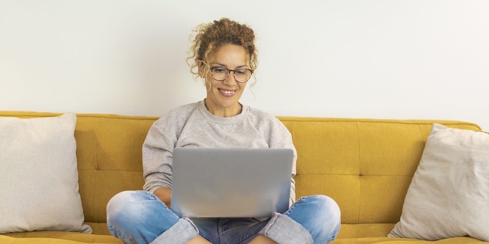 Happy Young Woman Working On Laptop Sitting On Sofa. Woman Wokring From Home. Young Woman In Eyeglas