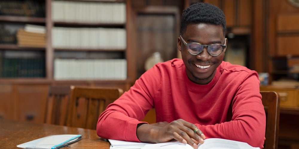 Happy young black student wearing spectacles reading text book. African american guy preparing for e
