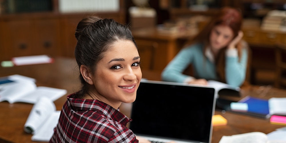 Happy young woman working on laptop and looking at camera. Portrait of smiling university student in