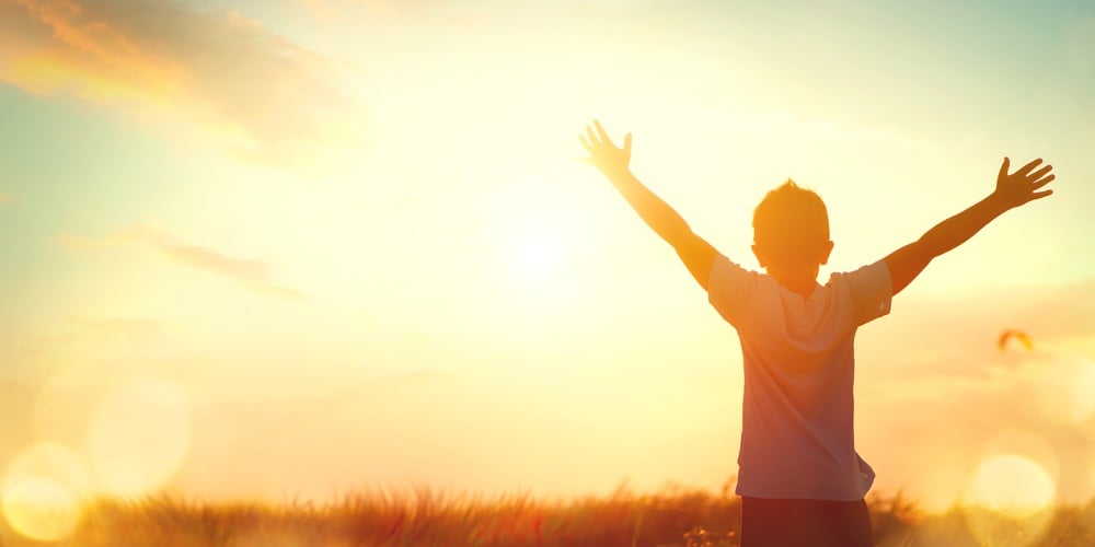 Little boy raising hands over sunset sky