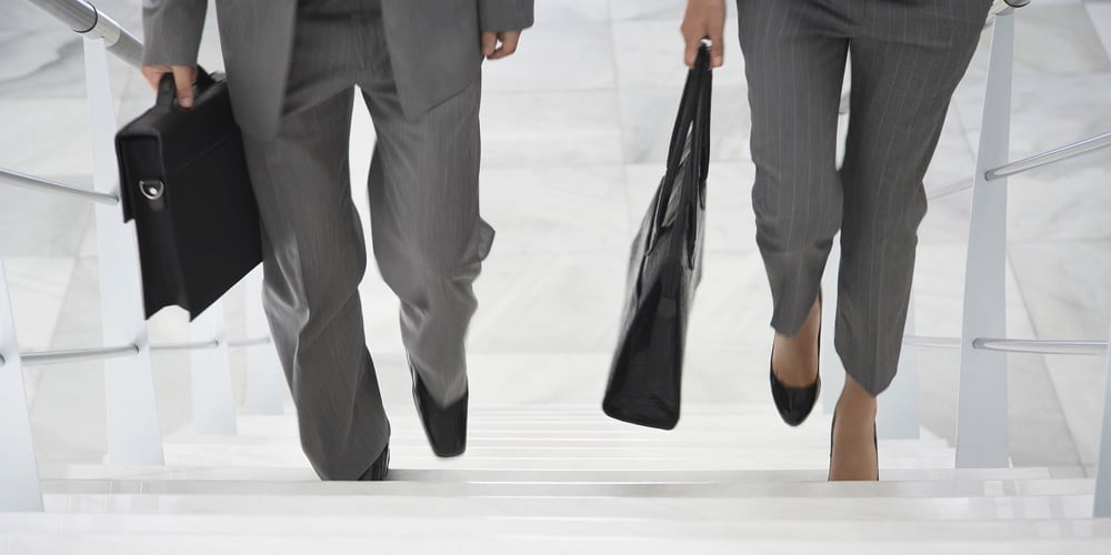 Lowsection of two businesspeople walking up stairs with bags in office