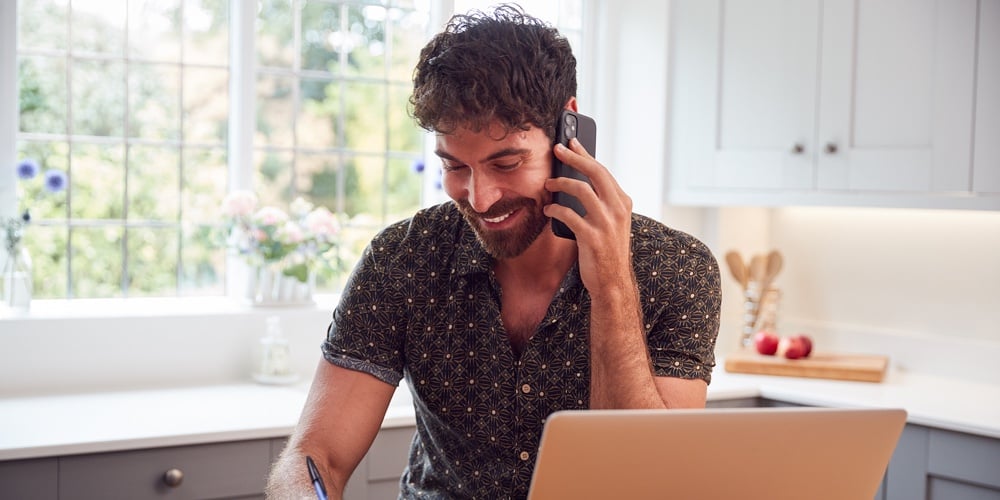 Man In Kitchen With Mobile Phone Working From Home Using Laptop During Health Pandemic