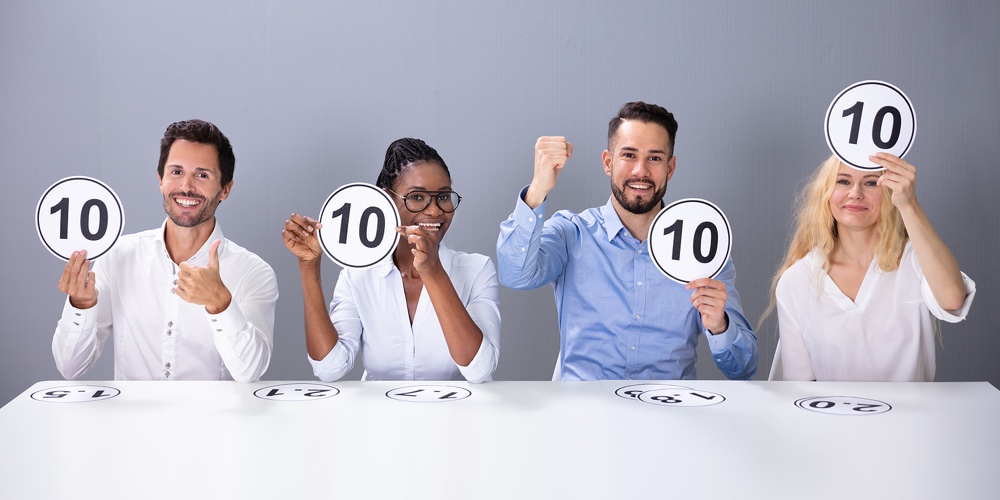 Panel Of Excited Judges Sitting In Front Of Grey Wall Showing 10 Score Cards