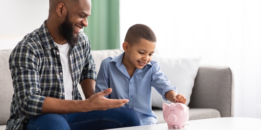 Personal Savings. African Father And Son Putting Coin In Piggybank Sitting On Sofa At Home