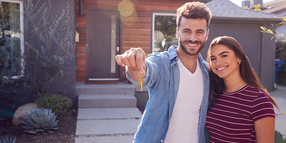 Portrait Of Couple Standing Outdoors In Front Of House With For Sale Sign In Garden Holding Keys