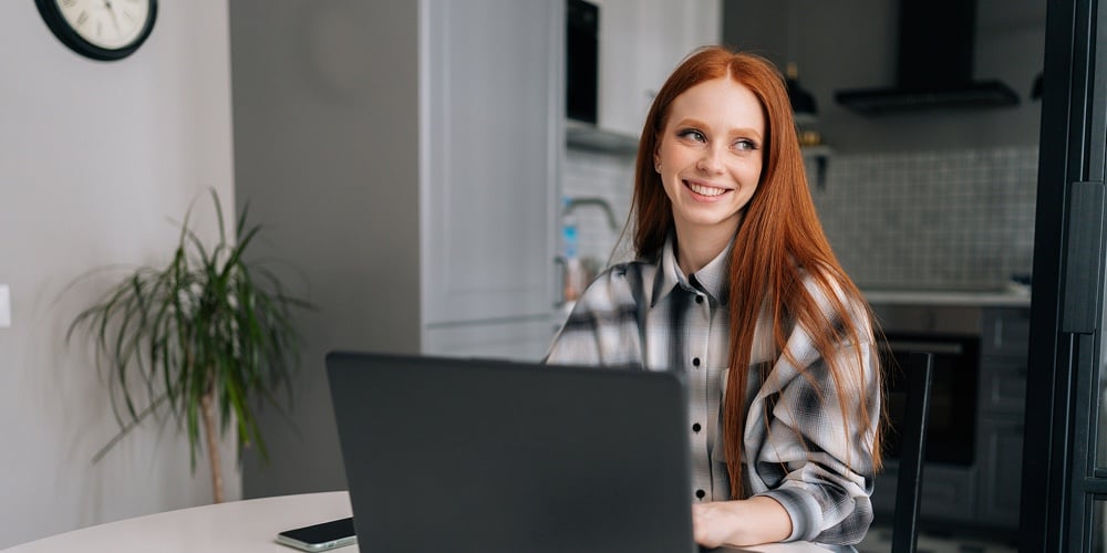 Portrait Of Happy Young Red-haired Woman Sitting At Desk With Laptop Computer And Mobile