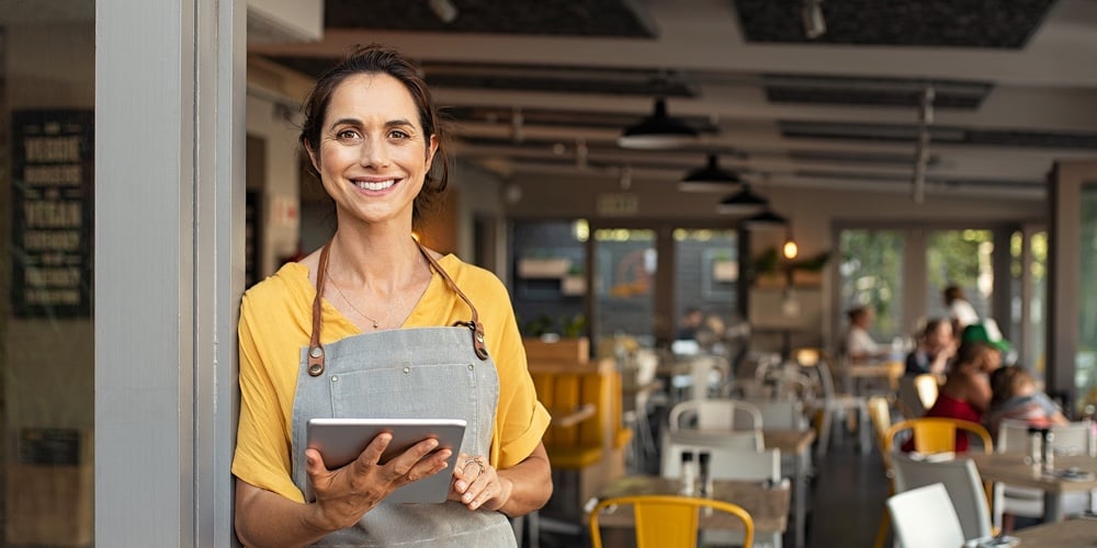 Portrait of happy woman standing at doorway of her store. Cheerful mature waitress waiting for clien