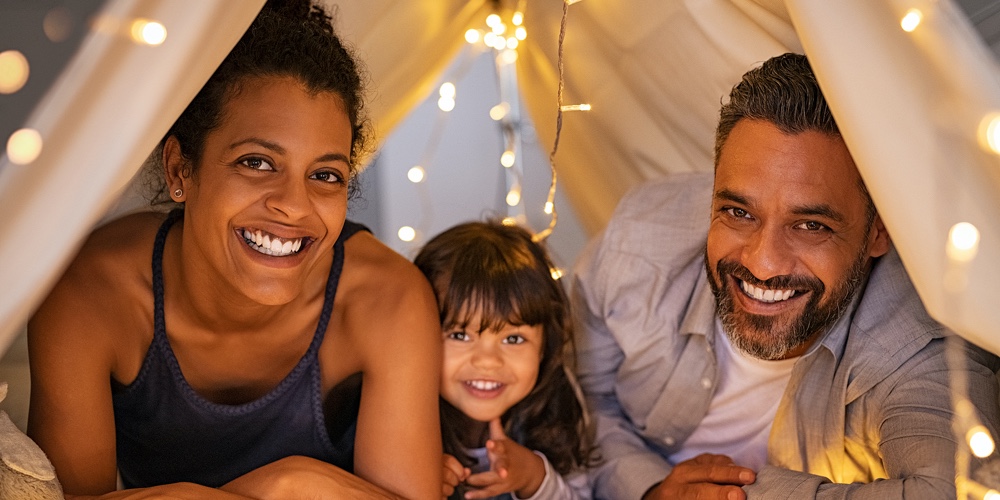 Portrait of little daughter lying with mother and father in illuminated tent. Cheerful man and happy