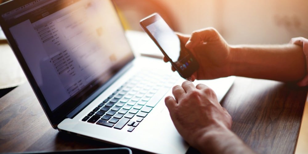Cropped image of a young male student texting on computer sitting at wooden table|Cropped shot of a young man working from home using smart phone and notebook computer