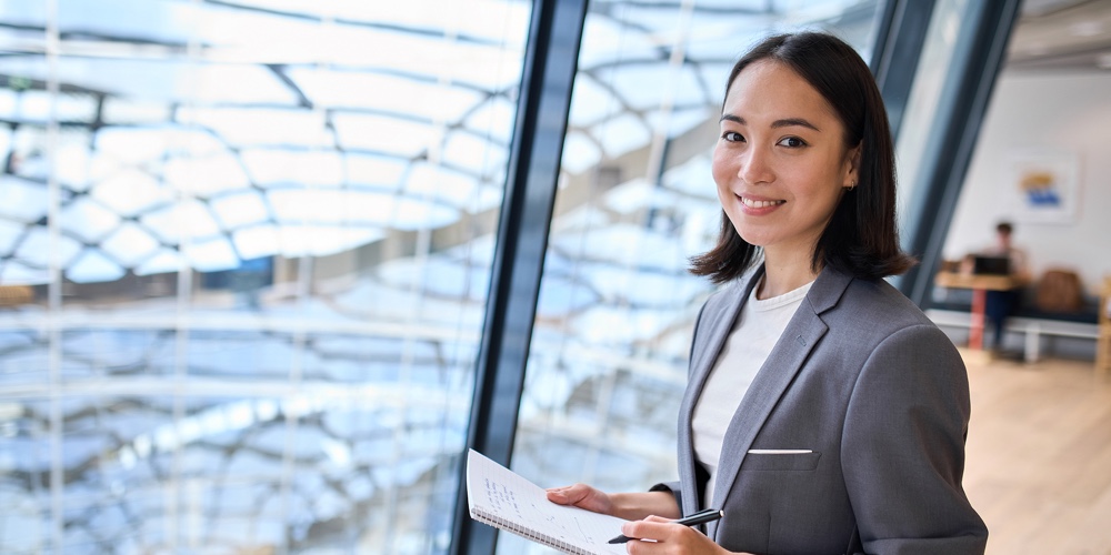 Smiling Young Asian Business Woman Manager Wearing Suit Holding Notebook Standing In Modern Glass Of