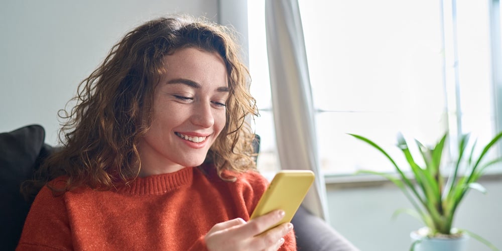 Smiling Young Woman Sitting On Couch Using Cell Phone