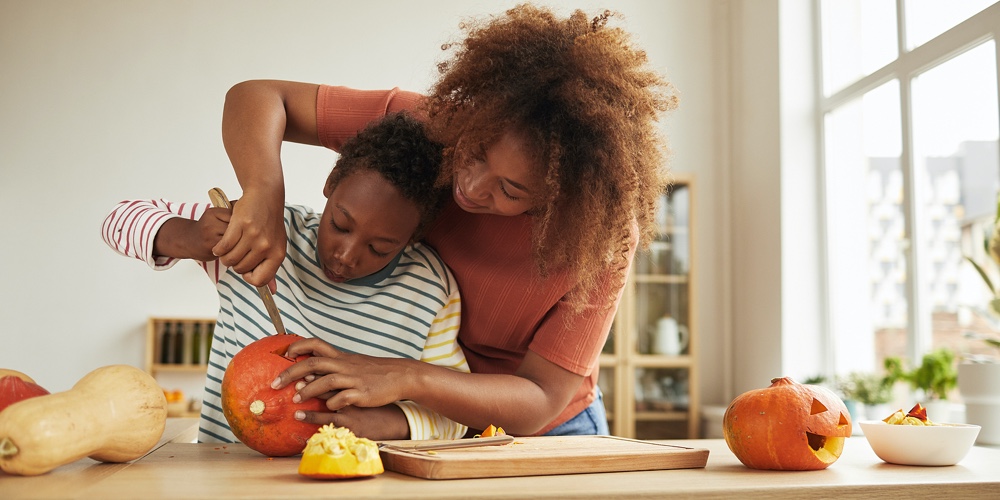 Stylish African American Woman Spending Time With Her Son Standing At Table Carving Pumpkin For Hall