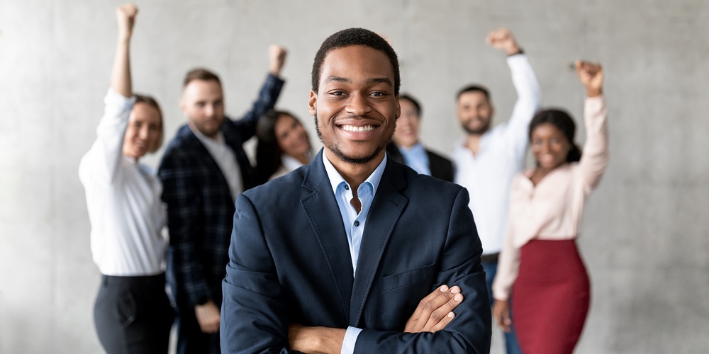 Successful Black Businessman Standing In Front Of Joyful Business Team In Modern Office, Smiling To