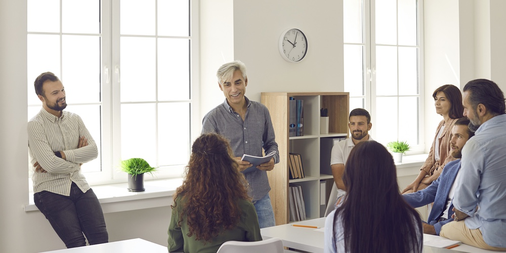 Team Of People Listening To Senior Business Coach During Training Meeting In Office