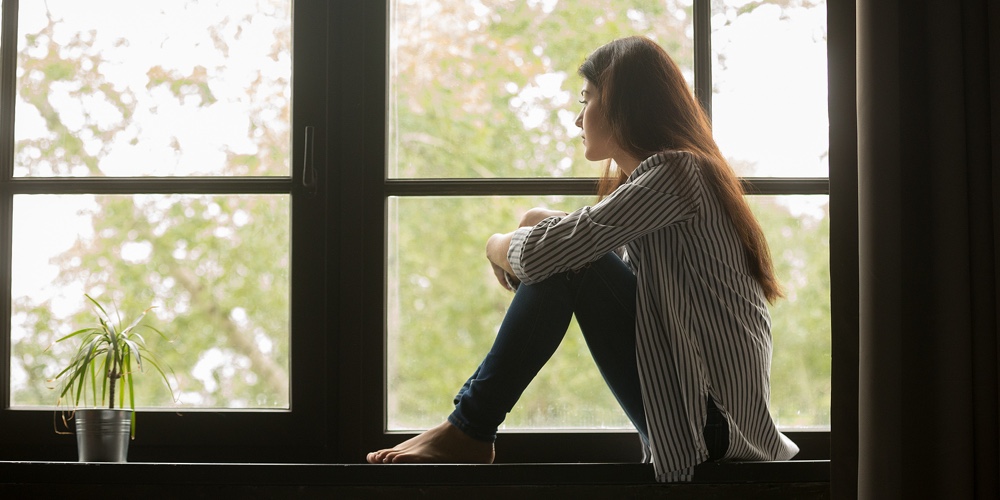 Thoughtful Girl Sitting On Sill Embracing Knees Looking At Window