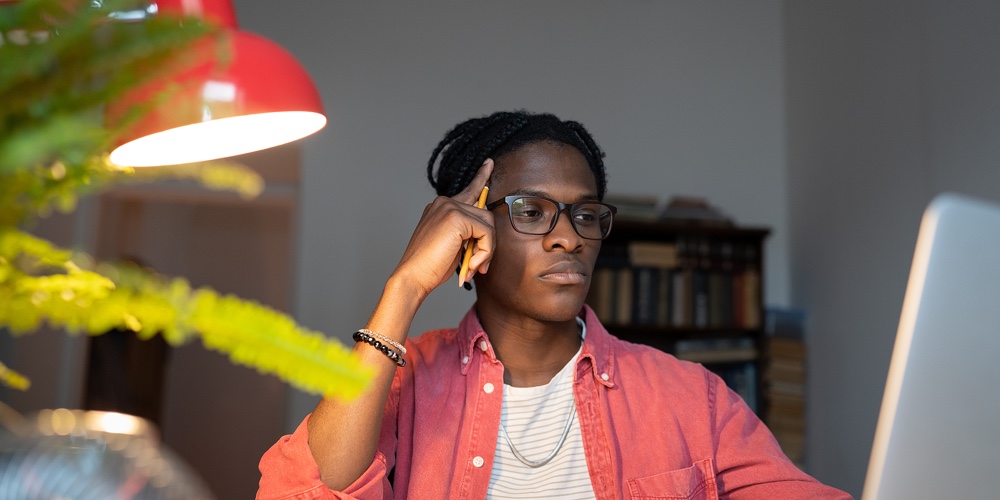 Tired Confused African American Man Looking At Laptop Screen Sits At Office Table With Notepad. Pens