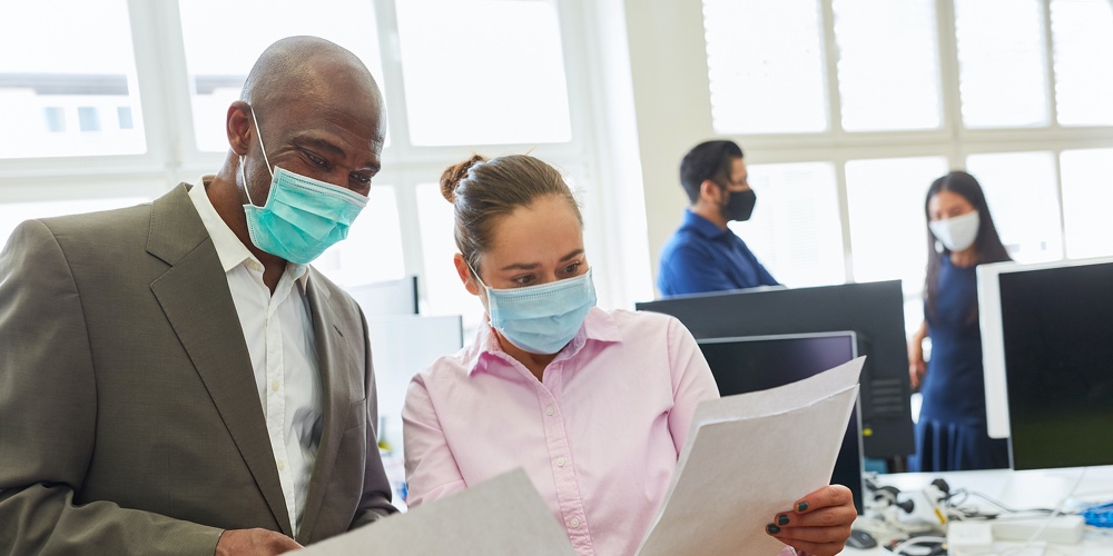 Two businessmen with face mask over Covid-19 talk about a business document in the office