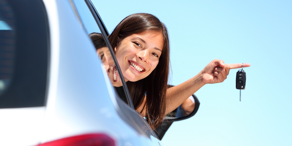 Woman driver holding car keys driving her new car. Beautiful multiracial young woman.