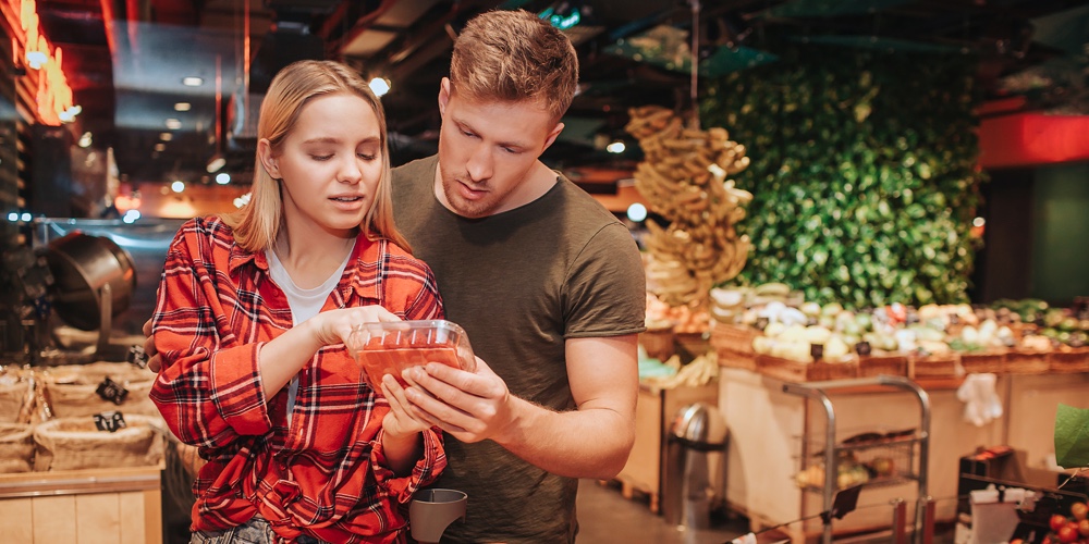 Young Couple In Grocery Store. They Stand Together And Read Text On Berries Plate. Trolley With Groc