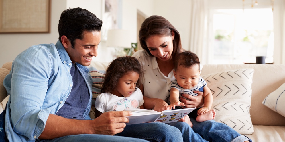 Young Hispanic family of four sitting on the sofa reading a book together in their living room