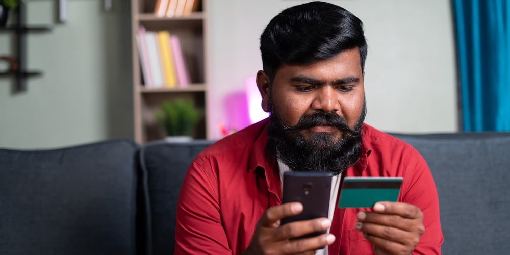 Young Man With Beard Making Online Payment Using Credit On Mobile Phone From Home – Concept Of Credi