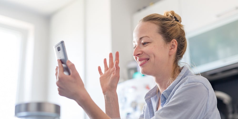 Young Smiling Cheerful Pleased Woman Indoors At Home Kitchen Using Social Media On Mobile Phone For