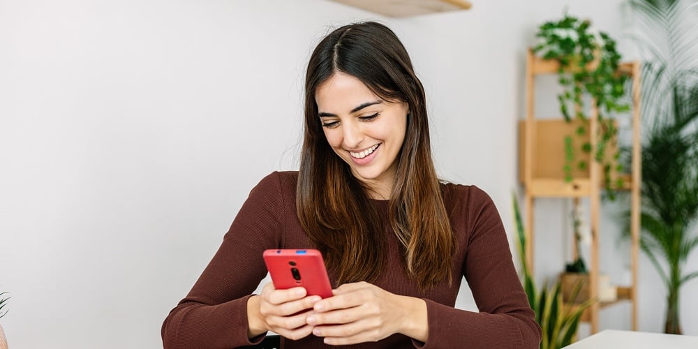 Young Smiling Happy Woman Using Mobile Phone While Working On Laptop At Home. Pretty Millennial Fema
