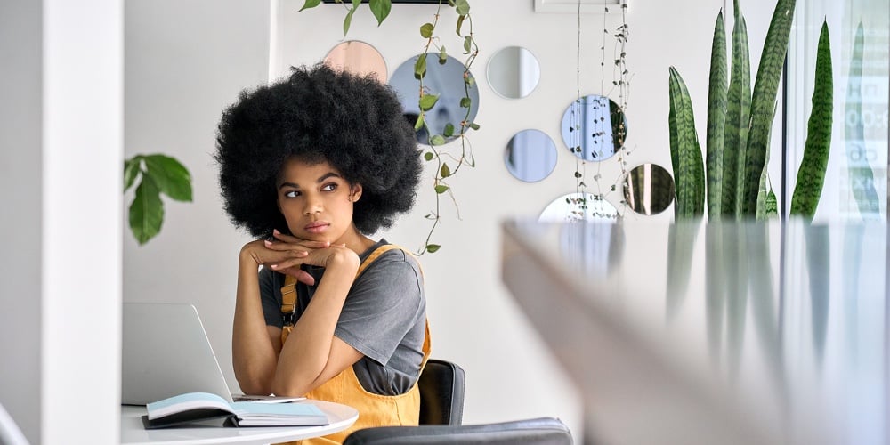 Young Thoughtful Dreamy African American Student Girl With Afro Hair Sitting At Table Holding Her Ch|Young Thoughtful Dreamy Smiling African American Student Generation Z Girl With Afro Hair Sitting At