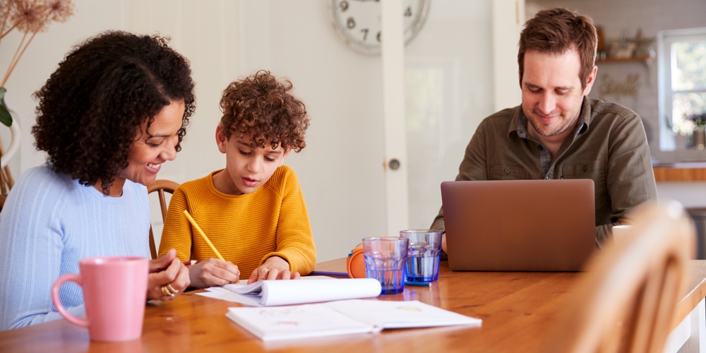 Father Works On Laptop As Mother Helps Son With Homework On Kitchen Table