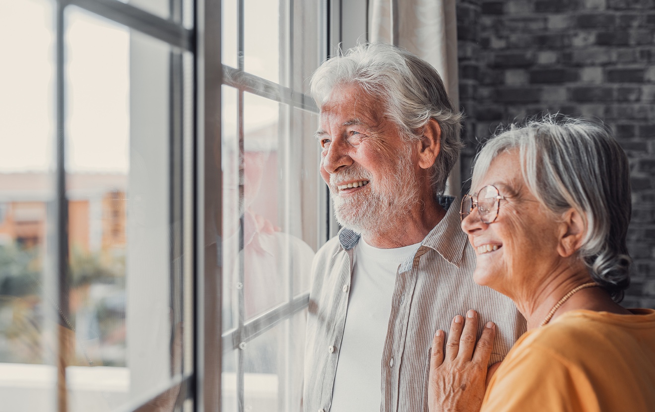 Happy Bonding Loving Middle Aged Senior Retired Couple Standing Near Window, Looking In Distance, Re