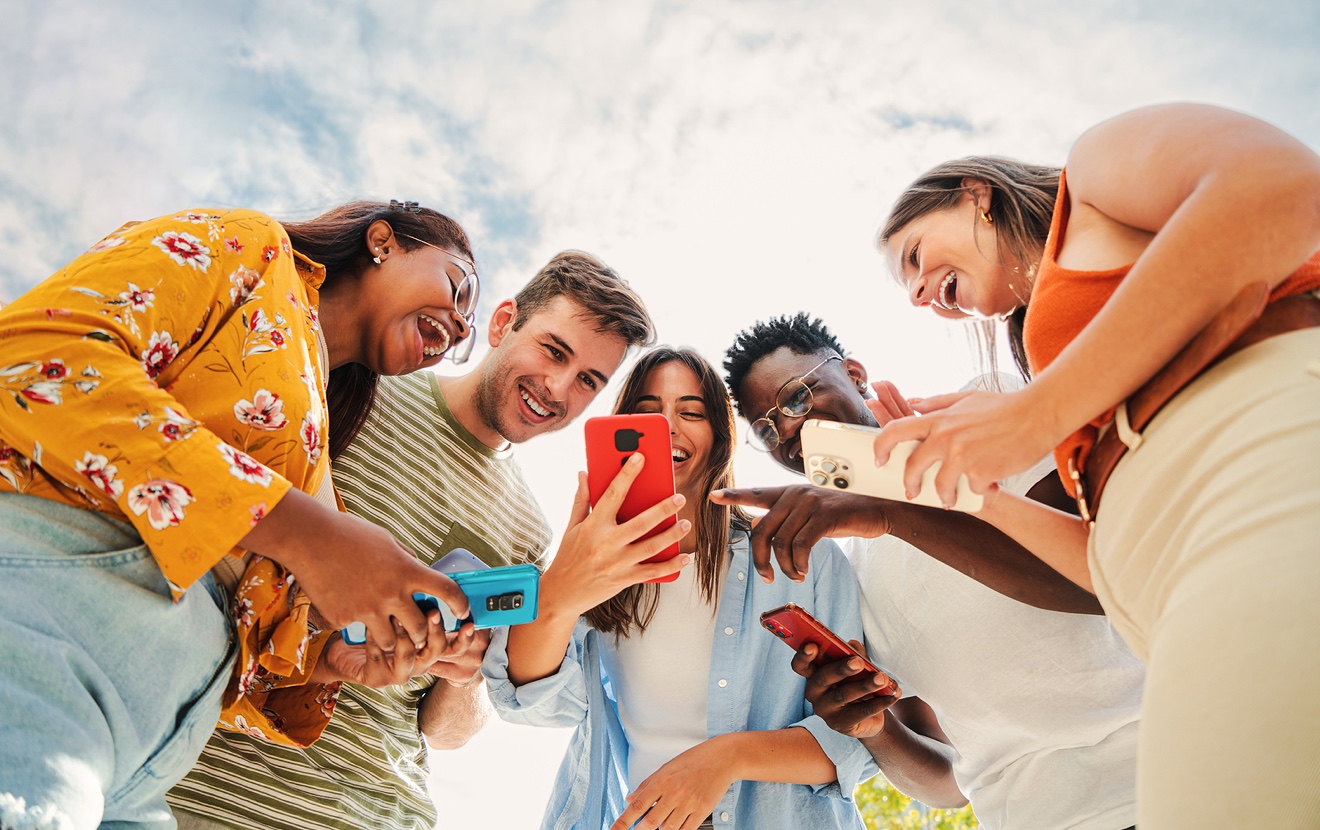 Low Angle View Of A Group Of Smiling Multiracial Teenagers Addicted To Smartphones, Watching Funny V