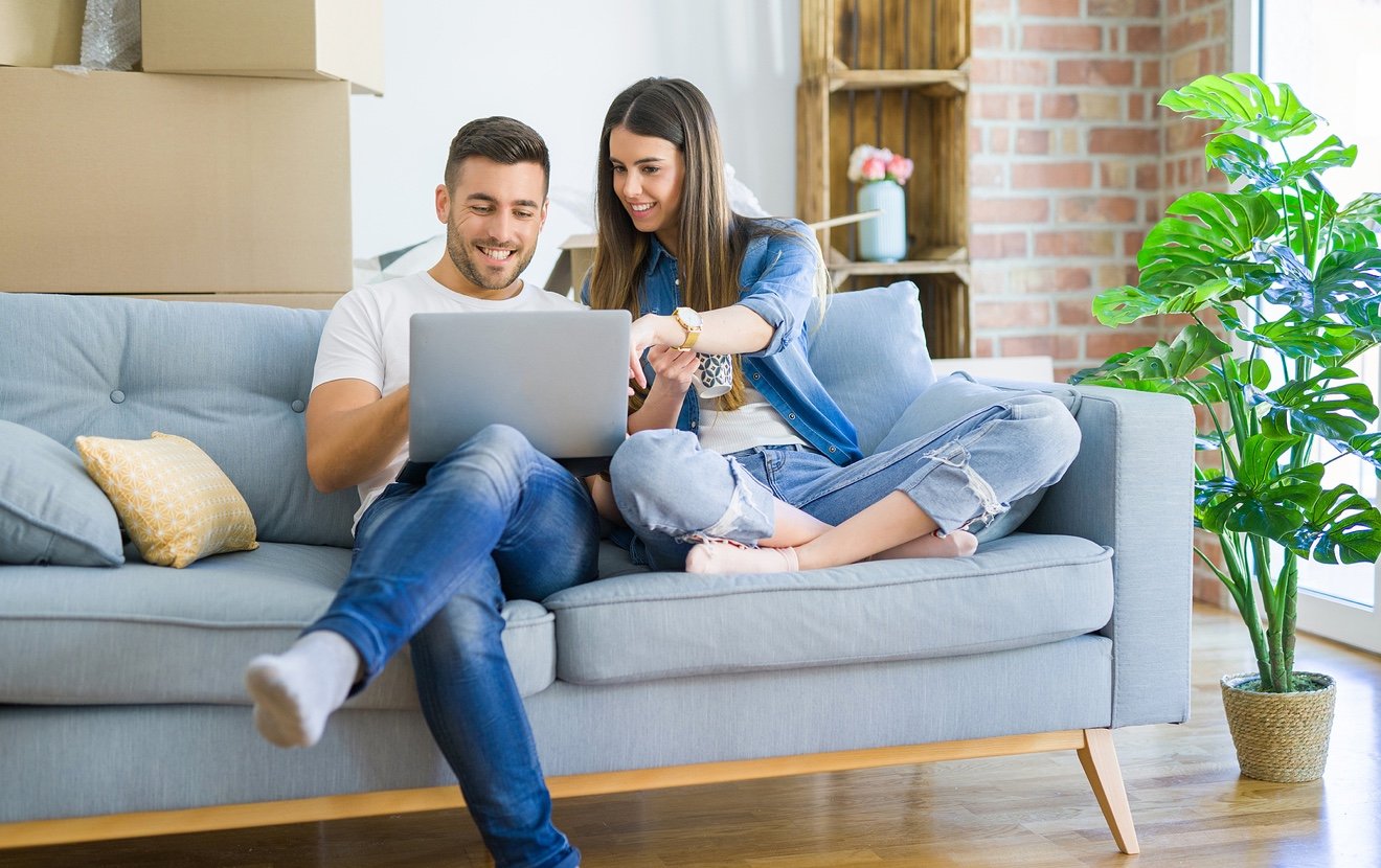 Young couple moving to a new home relaxing sitting on the sofa using computer laptop, smiling happy