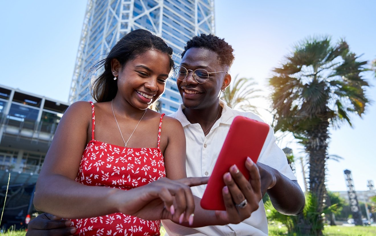 Young African American Couple Sitting Outdoors Using Smart Phone Together On Summer Holidays.