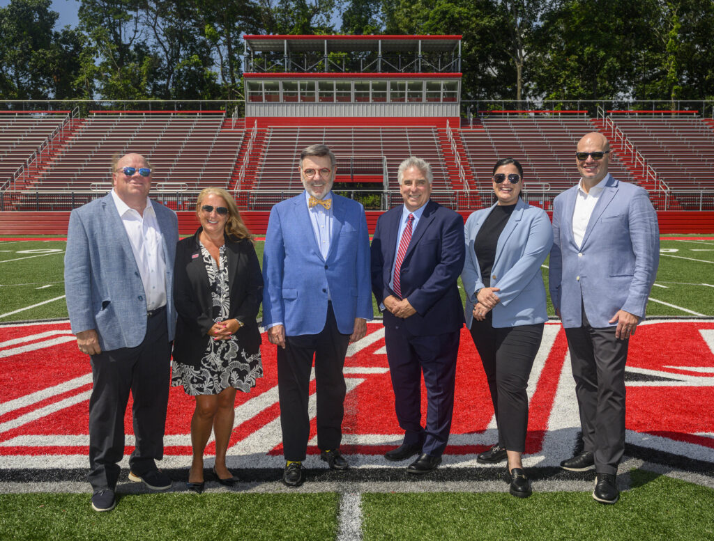 Six individuals pose on a football field with red bleachers in the background.